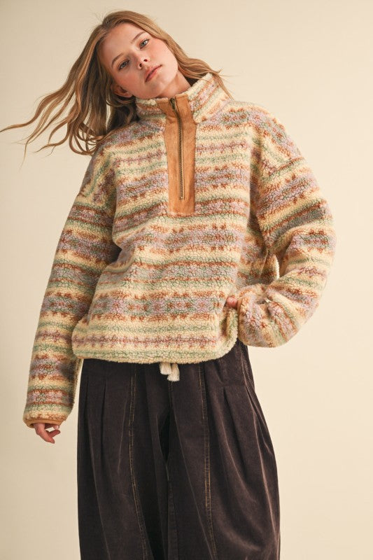 A woman wearing a fuzzy striped quarter-zip pullover sweater with horizontal bands of cream, tan, brown, and muted green, paired with dark brown corduroy pants, against a solid beige background.