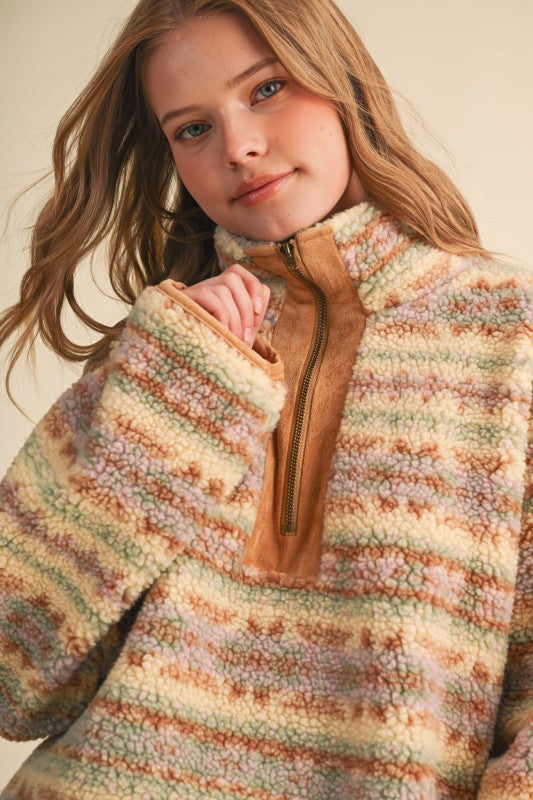 A close-up of a young woman wearing a fuzzy quarter-zip fleece pullover sweater with horizontal stripes in cream, tan, peach, and muted green, featuring a contrasting tan faux-leather placket and zipper.