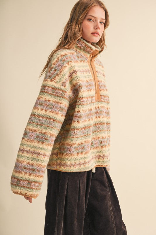 A woman wearing a fuzzy knit quarter-zip pullover sweater with horizontal multicolor stripes in beige, orange, green, and brown, paired with dark corduroy pants, set against a plain beige studio background.