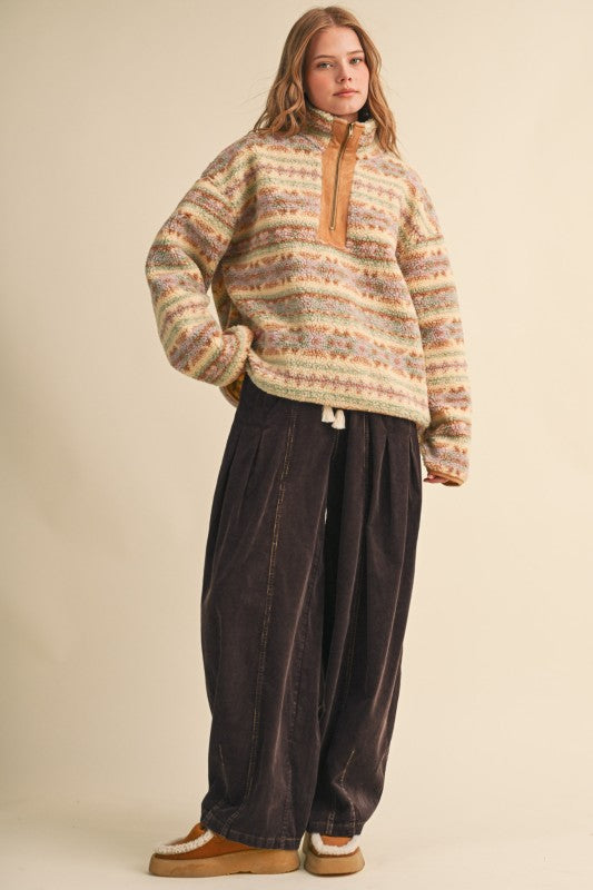A woman modeling a cozy, oversized quarter-zip knit sweater with horizontal multicolor stripes in beige, brown, and cream, paired with dark brown wide-leg pants and tan slip-on shoes, against a solid beige studio background.