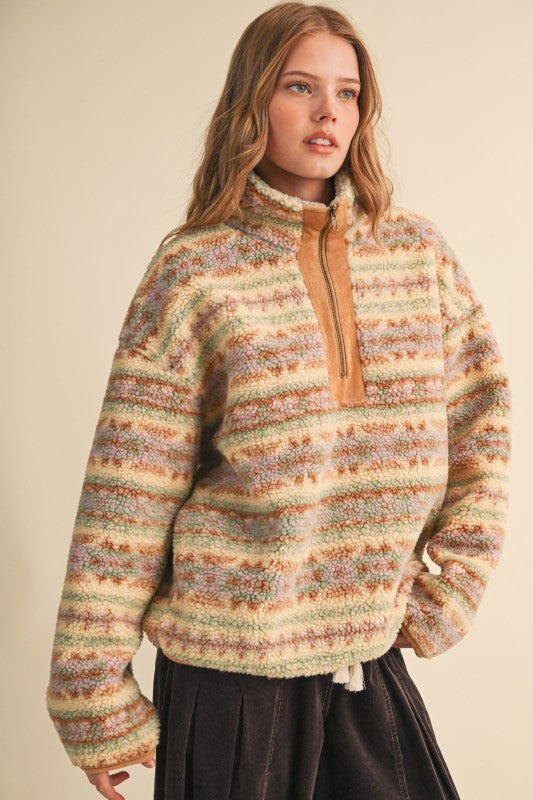 A woman wearing a fuzzy quarter-zip pullover sweater with horizontal multicolor stripes in beige, brown, green, and orange tones, paired with a dark skirt, set against a plain beige background.
